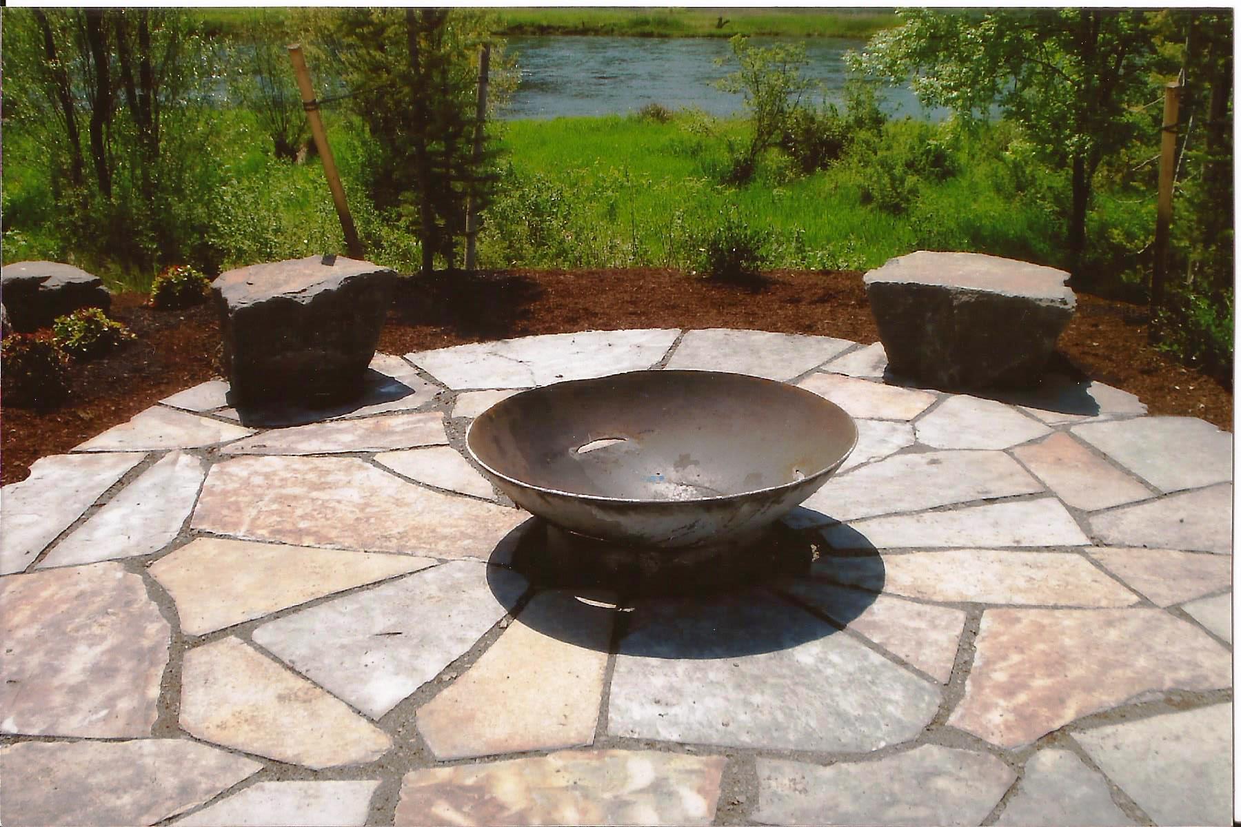 Image of a beautiful round paver patio with a fire pit in the middle, surrounded by a well-landscaped garden.