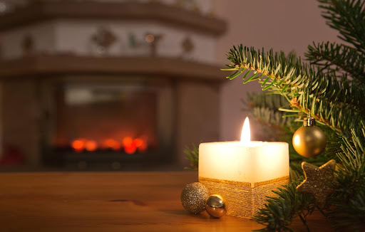 Image of a candle burning close to a Christmas tree with a burning fireplace in the background, a good reminder to consider holiday safety!